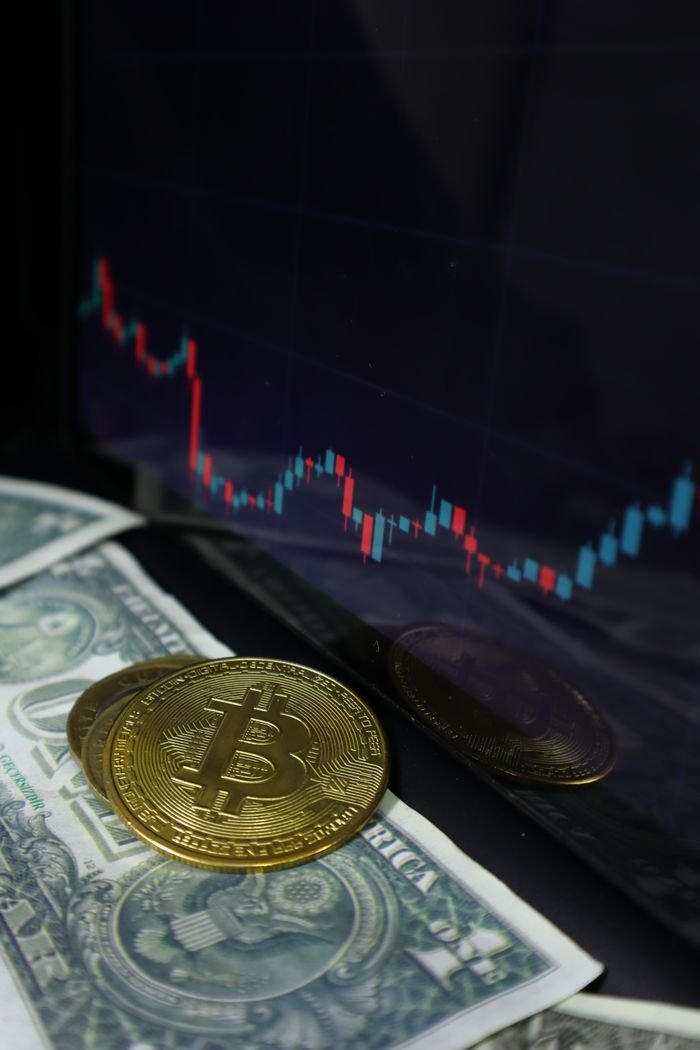 our-experience A close-up of Bitcoin coins with dollar bills and a stock market chart in the background.