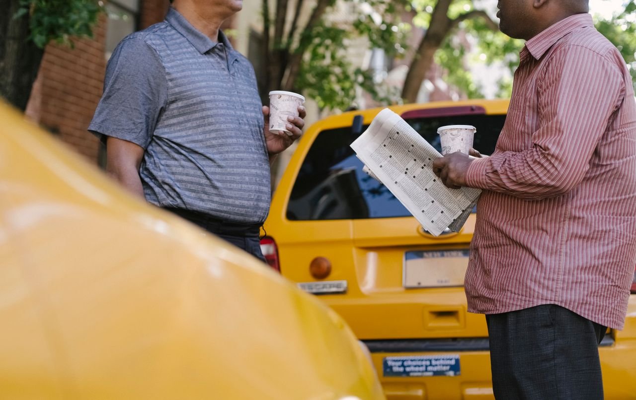 services-04 Crop anonymous multiracial taxi drivers with takeaway coffee and newspaper speaking near yellow cars in town